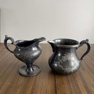 Könnte beinhalten: Zwei antike silberne Sahnekännchen. Das Kännchen links hat ein verziertes Blumendesign und einen Sockel. Das Kännchen rechts hat ein Blumendesign und einen gebogenen Griff. Beide Kännchen stehen auf einer Holzoberfläche.