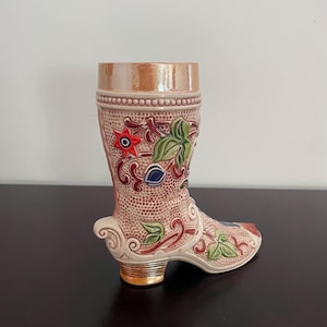 May include: A decorative ceramic boot-shaped vase. The boot is primarily a light brown color with intricate floral and leaf designs in red, green, and blue. The heel and top rim have a gold metallic finish. The boot has a textured surface.