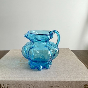 May include: A turquoise glass creamer with a bulbous body and a handle. The creamer has a ruffled rim and sits on a textured surface. The creamer is a decorative item.
