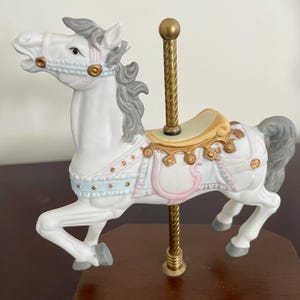 May include: A white carousel horse figurine with gray mane and tail, adorned with light blue and pink accents. The horse has a gold-colored pole and saddle, and is mounted on a wooden base. A decorative piece.
