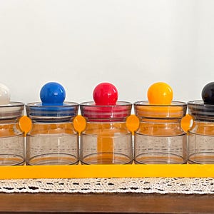 May include: A set of six clear glass spice jars with colorful lids. The lids are white, blue, red, yellow, black, and gray. The jars are sitting on a yellow rack.
