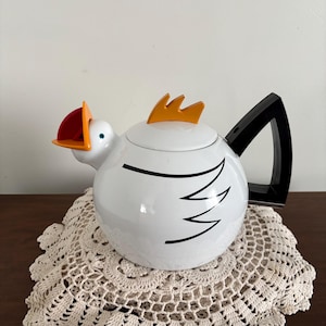 May include: A white teapot shaped like a chicken with a black angular handle, an orange crest on the lid, and an orange and red beak. The body features a black line drawing of a wing, and the spout is integrated into the chicken's head design.
