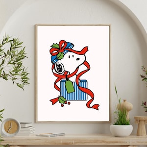 Op de afbeelding: Een ingelijste print met Snoopy uit Peanuts, verpakt in een rood lint en een blauwe geschenkdoos. Het kunstwerk bevat hulst en een groen label, ideaal voor feestelijke decoratie. De print wordt tentoongesteld op een houten tafel.