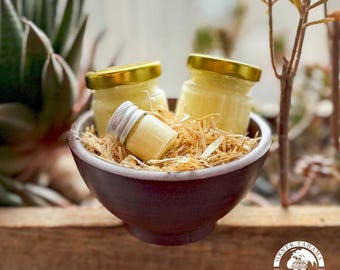 May include: Three small glass jars filled with a pale yellow cream, nestled in a brown bowl lined with straw. The jars have gold lids and are arranged in a triangular shape. The bowl is sitting on a wooden surface.