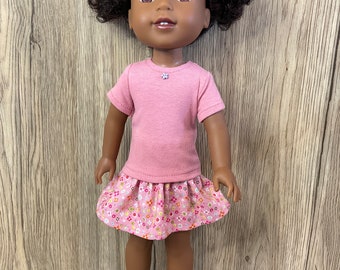 Puede incluir: Una muñeca con piel oscura y cabello negro rizado peinado en dos coletas. La muñeca viste una camiseta rosa y una falda floral rosa. Zapatos blancos con lazos completan el atuendo. Una pequeña flor está en la camisa.
