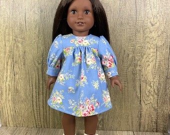 May include: A doll in a light blue floral dress featuring pink and white roses. The dress has long sleeves and a gathered neckline. The doll is wearing white shoes with a strap.