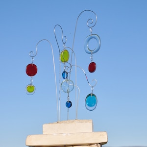 May include: A metal sculpture with colorful glass circles hanging from it. The sculpture is on a white base and is against a blue sky.