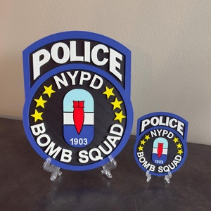 May include: Two NYPD Bomb Squad badges, one larger than the other, are displayed on a wooden stand. Both badges are blue with white lettering and a yellow star design. The smaller badge is a miniature version of the larger badge.
