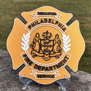 May include: A gold-colored, 3D printed, fire department badge with the words "Philadelphia Fire Department Service" and "Dedication" around the perimeter. The badge features a black and white crest with the city's seal in the center.
