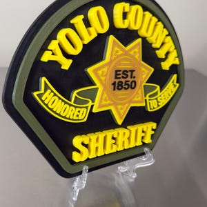 Peut inclure: Un &eacute;cusson de sh&eacute;rif du comt&eacute; de Yolo noir, jaune et vert. L'&eacute;cusson comporte les mots "YOLO COUNTY SHERIFF", "HONORED" et "TO SERVE". Le centre de l'&eacute;cusson comporte une &eacute;toile avec le texte "EST. 1850".