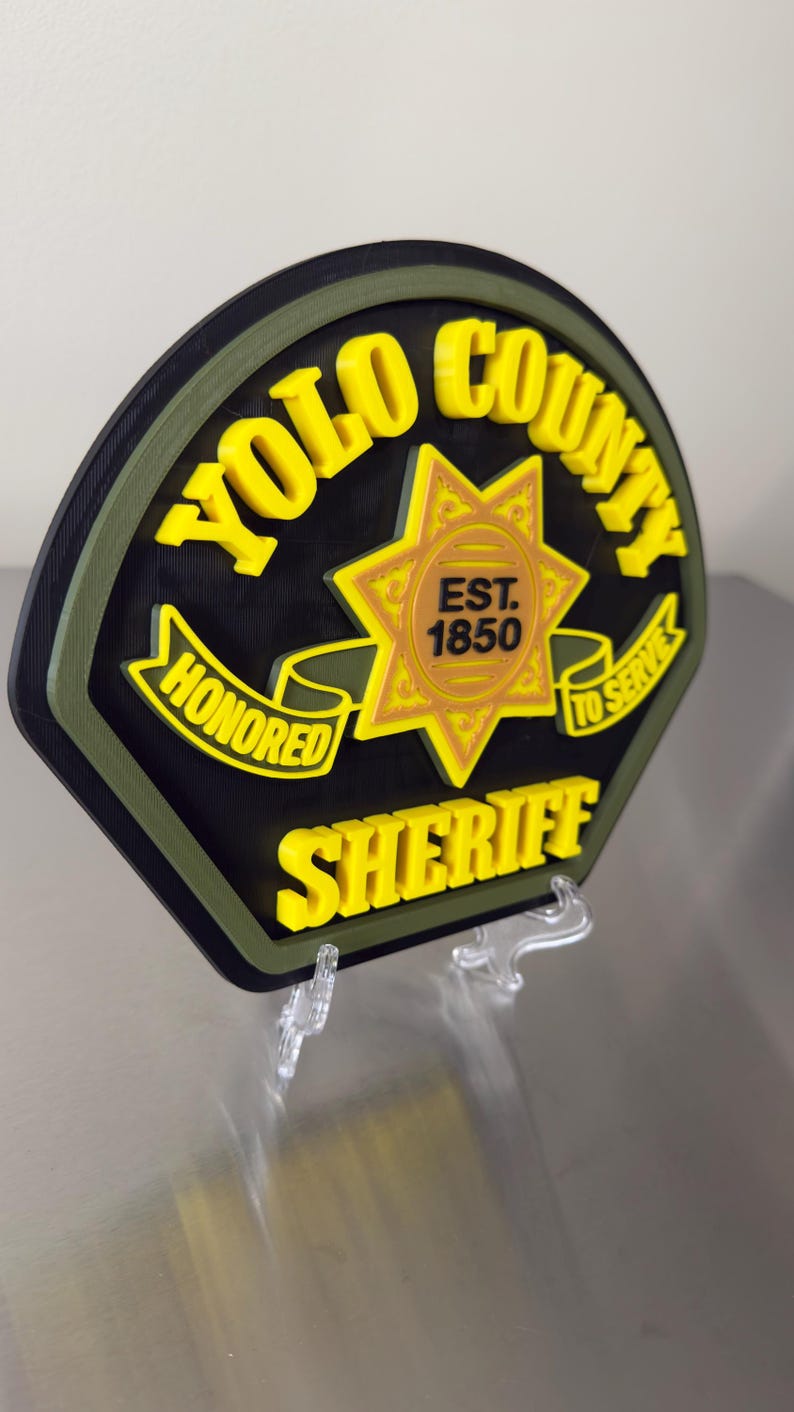 Peut inclure: Un &eacute;cusson de sh&eacute;rif du comt&eacute; de Yolo noir, jaune et vert. L'&eacute;cusson comporte les mots "YOLO COUNTY SHERIFF", "HONORED" et "TO SERVE". Le centre de l'&eacute;cusson comporte une &eacute;toile avec le texte "EST. 1850".