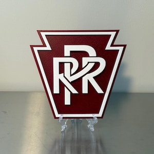 May include: A maroon and white sign featuring the letters "RR" in a stylized design. The sign is in the shape of a keystone, with a white border. The sign is displayed on a clear plastic stand.