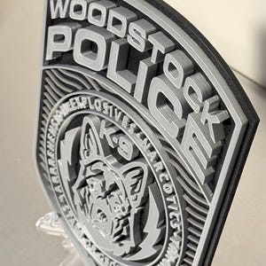 May include: A 3D-printed Woodstock Police badge with raised lettering and a K-9 unit emblem. The badge features the words "Woodstock Police" and "K-9 Explosives Narcotics" in gray against a black border. The badge is displayed on a clear stand.