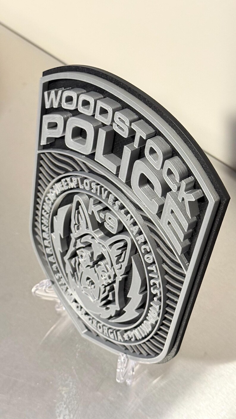 May include: A 3D-printed Woodstock Police badge with raised lettering and a K-9 unit emblem. The badge features the words "Woodstock Police" and "K-9 Explosives Narcotics" in gray against a black border. The badge is displayed on a clear stand.