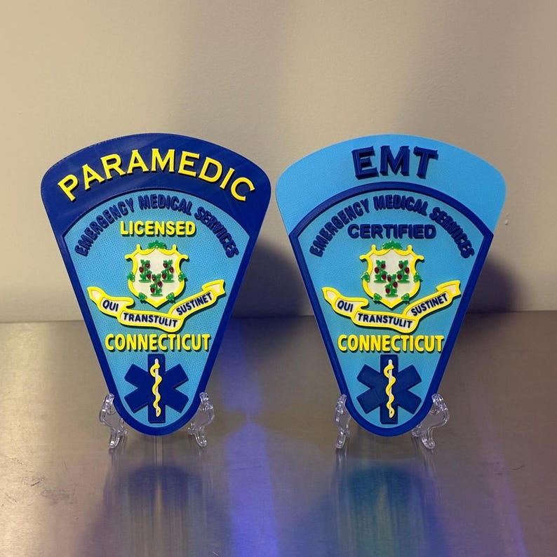 Op de afbeelding: Twee blauwe en gele badges van de ambulancedienst. E&eacute;n leest "PARAMEDIC" en "LICENSED", de andere leest "EMT" en "CERTIFIED". Beide hebben de tekst "EMERGENCY MEDICAL SERVICES", "CONNECTICUT" en een life star symbool.