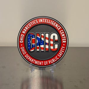 May include: A round emblem with the text "OHIO NARCOTICS INTELLIGENCE CENTER" in red and white. The center features the letters "ONIC" with the Ohio state flag design. The emblem is displayed on a clear stand.