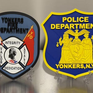 Op de afbeelding: Twee decoratieve plaquettes: een voor de brandweer van Yonkers en een voor de politie van Yonkers. De brandweerplaquette is blauw met een Amerikaanse vlag en de woorden "Integriteit, Moed, Toewijding, Loyaliteit". De politieplaquette is blauw en geel.