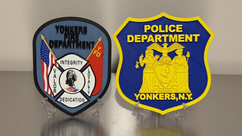 Op de afbeelding: Twee decoratieve plaquettes: een voor de brandweer van Yonkers en een voor de politie van Yonkers. De brandweerplaquette is blauw met een Amerikaanse vlag en de woorden "Integriteit, Moed, Toewijding, Loyaliteit". De politieplaquette is blauw en geel.