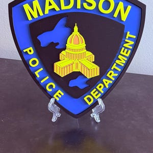 May include: A shield-shaped emblem for the Madison Police Department. The design features the city name in yellow, a state outline, and a building illustration. The emblem is black with a blue border.