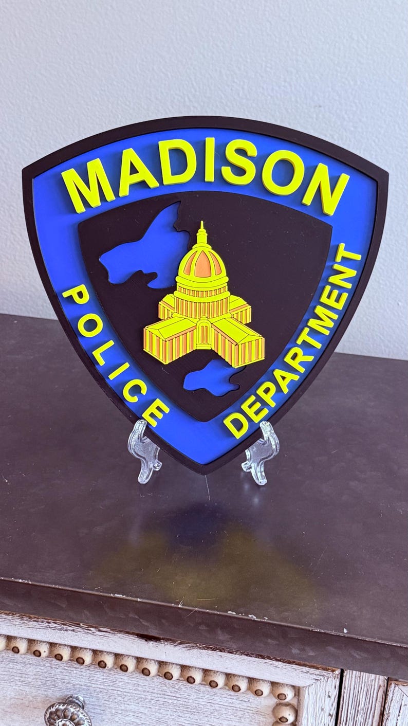 May include: A shield-shaped emblem for the Madison Police Department. The design features the city name in yellow, a state outline, and a building illustration. The emblem is black with a blue border.