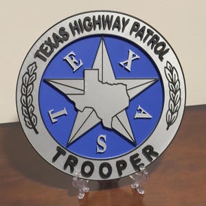 May include: A circular Texas Highway Patrol Trooper emblem featuring a blue center with a silver star and a silhouette of the state of Texas. The border is silver with black lettering and two wheat stalks. The letters T, E, X, A, S are placed around the star.