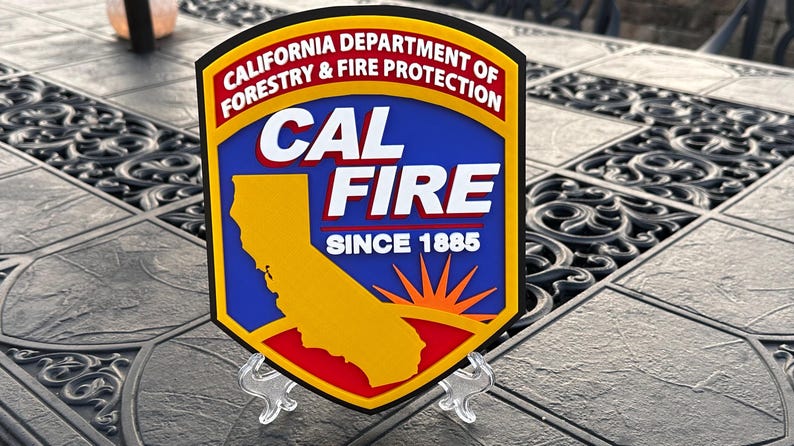May include: A shield-shaped emblem for the California Department of Forestry & Fire Protection. The emblem features a yellow outline, a blue center with the words "CAL FIRE" in white, and a yellow California map. The text "SINCE 1885" is also present.