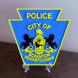 May include: A blue and yellow keystone-shaped sign featuring the text POLICE CITY OF SCRANTON PENNSYLVANIA in raised yellow lettering. The center displays a yellow emblem with two black horses flanking a central crest with a bird icon.