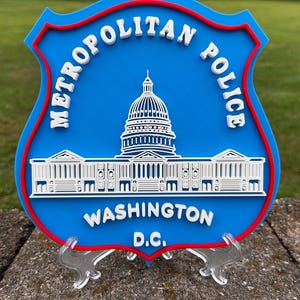May include: A shield-shaped Metropolitan Police Washington D.C. sign featuring a blue background with a red border. The sign displays a white raised graphic of the United States Capitol building and white text reading Metropolitan Police Washington D.C.