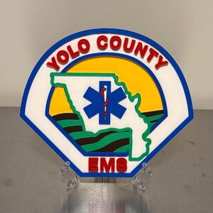 May include: A colourful emblem featuring the words "YOLO COUNTY" in red, arched over a yellow circle. The emblem includes a blue star of life symbol and the word "EMS" in red. The design is set against a white and blue background.