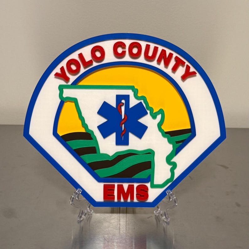 May include: A colourful emblem featuring the words "YOLO COUNTY" in red, arched over a yellow circle. The emblem includes a blue star of life symbol and the word "EMS" in red. The design is set against a white and blue background.