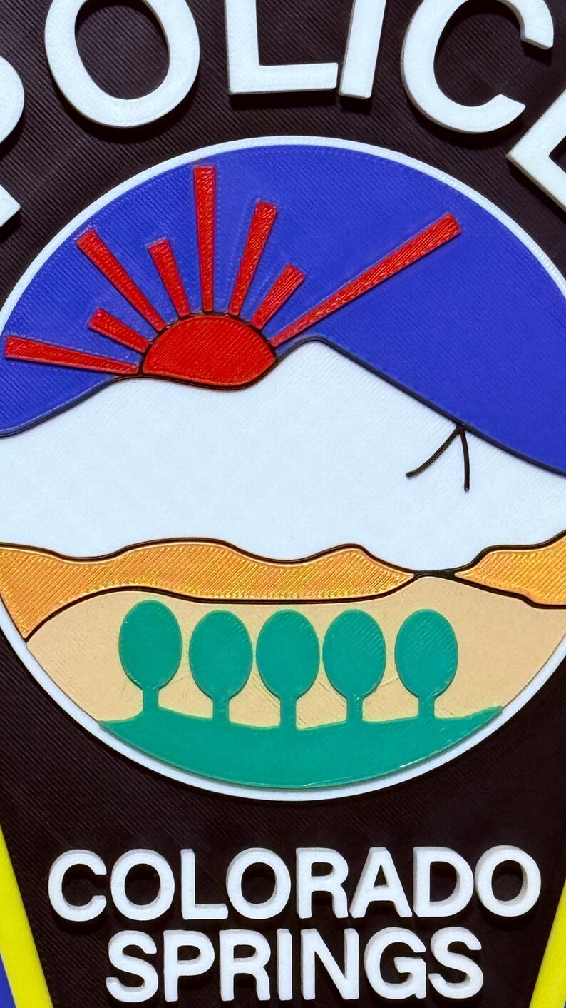 K&ouml;nnte beinhalten: Ein schwarz-wei&szlig;es Polizeibadge mit einer blauen, roten und wei&szlig;en Bergszene mit einer roten Sonne und f&uuml;nf gr&uuml;nen B&auml;umen. Der Text "COLORADO SPRINGS" ist in wei&szlig;en Buchstaben unter der Szene geschrieben.