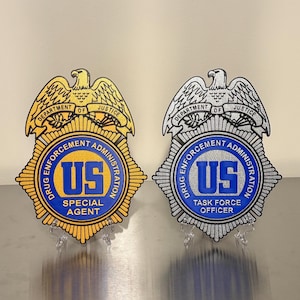 May include: Two badges with the text "DRUG ENFORCEMENT ADMINISTRATION" and "DEPARTMENT OF JUSTICE". One badge reads "SPECIAL AGENT" and the other reads "TASK FORCE OFFICER". The badges are gold and silver with blue and yellow accents.
