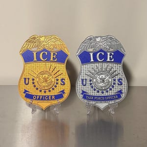 May include: Two shield-shaped badges featuring an eagle at the top. The left badge is gold with blue accents, reading ICE U.S. OFFICER. The right badge is silver with blue accents, reading ICE U.S. TASK FORCE OFFICER. Both include the DHS seal.