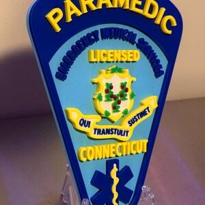 Op de afbeelding: Een blauw en geel Paramedic-bord met de tekst "Emergency Medical Services" en "Licensed". Het bord toont het zegel van de staat Connecticut en een blauw symbool van de levensster.