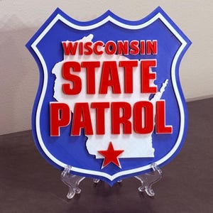 May include: A blue, white, and red wooden sign featuring the words "WISCONSIN STATE PATROL" in bold letters. The sign is shaped like a shield with a white outline and a red star at the bottom. The state's outline is in white.