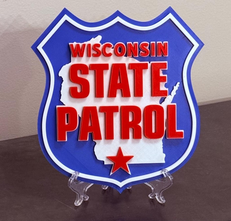 May include: A blue, white, and red wooden sign featuring the words "WISCONSIN STATE PATROL" in bold letters. The sign is shaped like a shield with a white outline and a red star at the bottom. The state's outline is in white.