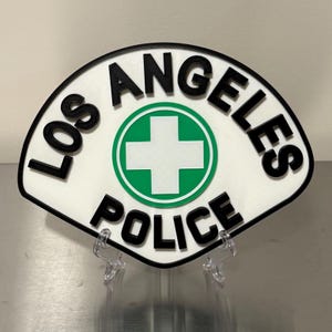 May include: A fan-shaped sign with the words "LOS ANGELES POLICE" in black, bold letters. The sign has a white background with a green circle and a white cross in the center. The sign is displayed on a clear stand.