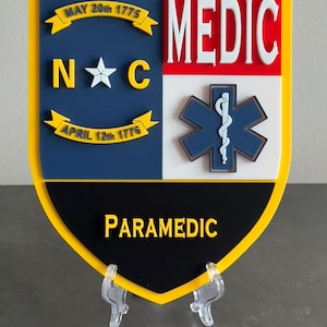 May include: A shield-shaped emblem with a yellow border, featuring the words "MEDIC" in white on a red background. The emblem includes a blue and white star of life symbol, and the word "PARAMEDIC" in yellow. The top left section has the text "MAY 20th 1775".