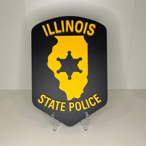 May include: A black and yellow Illinois State Police badge. The badge features the state outline in yellow with a star in the center. The words "ILLINOIS" and "STATE POLICE" are in yellow.