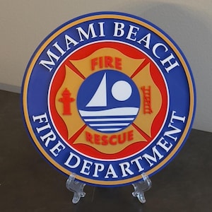 May include: A blue and gold round plaque with the words "Miami Beach Fire Department" around the edge. The center features a red and gold fire department symbol with a boat, a fire hydrant, and a ladder.