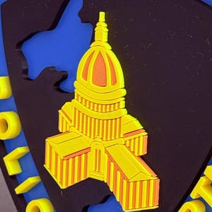 May include: A yellow and orange emblem featuring a building design, likely a capitol building, against a black and blue background. The word "MADISON" is at the top in yellow letters, and "POLICE DEPART" is at the bottom.