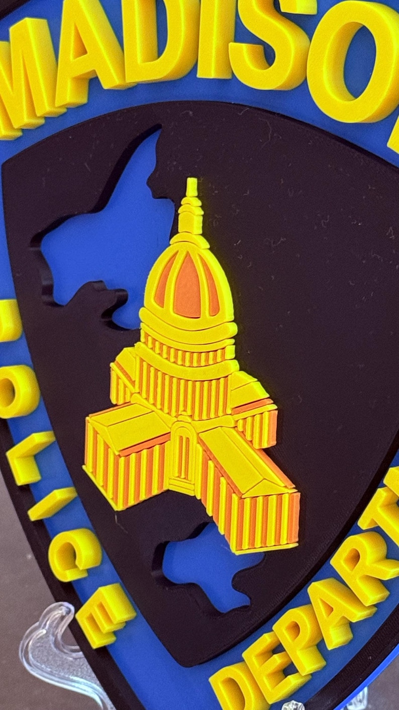 May include: A yellow and orange emblem featuring a building design, likely a capitol building, against a black and blue background. The word "MADISON" is at the top in yellow letters, and "POLICE DEPART" is at the bottom.