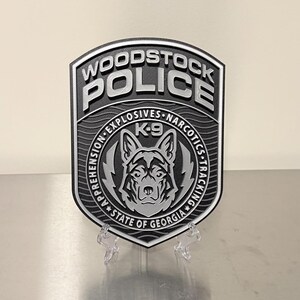 May include: A black and silver Woodstock Police badge with the words "WOODSTOCK POLICE" at the top. The badge features a K-9 unit emblem with the text "EXPLOSIVES NARCOTICS TRACKING" and "STATE OF GEORGIA".