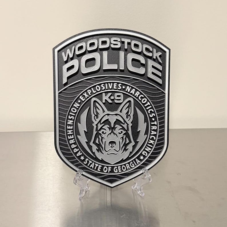 May include: A black and silver Woodstock Police badge with the words "WOODSTOCK POLICE" at the top. The badge features a K-9 unit emblem with the text "EXPLOSIVES NARCOTICS TRACKING" and "STATE OF GEORGIA".
