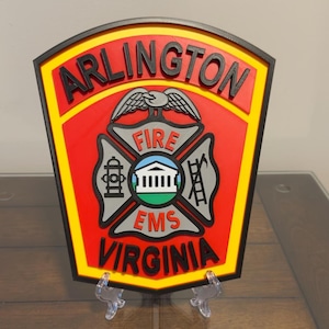 May include: A multi-layered wall plaque featuring the Arlington Fire EMS Virginia emblem. The shield-shaped design includes black, red, yellow, and grey colors with raised lettering and a central crest depicting a building, a fire hydrant, and a ladder.