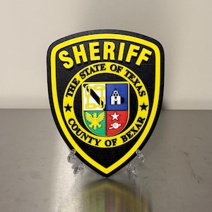 May include: A black and yellow shield-shaped emblem with the word "SHERIFF" at the top. The emblem features the text "THE STATE OF TEXAS" and "COUNTY OF BEXAR". The center has a colorful crest design.