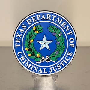May include: A circular emblem for the Texas Department of Criminal Justice. The design features a blue background with a white star in the center, surrounded by a wreath and the words "TEXAS DEPARTMENT OF CRIMINAL JUSTICE". The emblem is displayed on a clear stand.