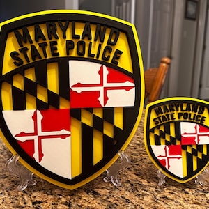 May include: Two shield-shaped Maryland State Police emblems in different sizes, featuring the Maryland state flag design in yellow, black, white, and red, with the text MARYLAND STATE POLICE in black lettering at the top of each emblem.