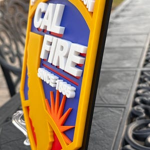 May include: A 3D sign featuring the text "CAL FIRE" in white, with a yellow border and a blue background. The sign also includes the text "SINCE 1885" and a graphic of the state of California. The sign is mounted on a clear stand.