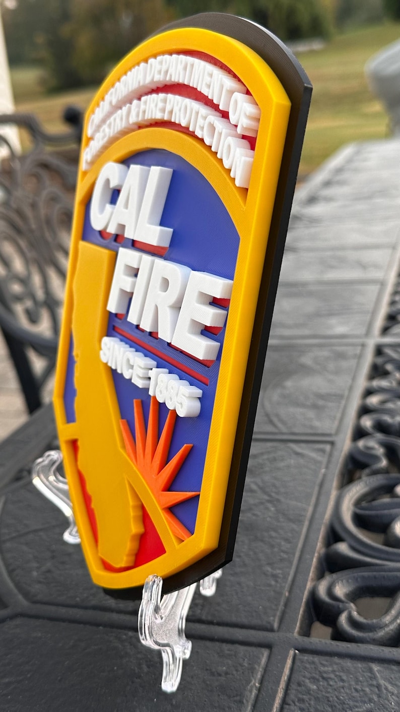 May include: A 3D sign featuring the text "CAL FIRE" in white, with a yellow border and a blue background. The sign also includes the text "SINCE 1885" and a graphic of the state of California. The sign is mounted on a clear stand.
